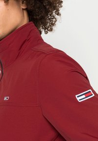 Rote technische Jacke mit hohem Kragen. Verfügt über ein Logo-Patch am linken Ärmel, glatte Textur und Nahtdetails.