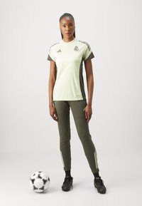 Maglietta sportiva verde chiaro con accenti verde scuro, distintivo sul petto; abbinata a pantaloni verde scuro con strisce gialle e scarpette nere.