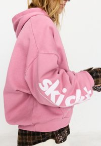 Rózsaszín kapucnis pulóver puha anyagból, nagy fehér "ski club" felirattal az ujjon. A kialakítás laza szabást és kapucnit tartalmaz.