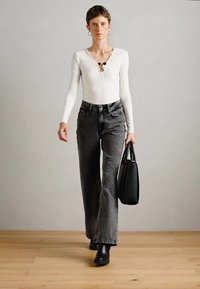White long-sleeve top with a gathered detail and keyhole; paired with high-waisted, flared dark gray jeans and black ankle boots, holding a black bag.
