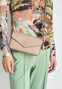 Béžová crossbody taška vyrobená z texturovaného materiálu, s klopou a stříbrnými doplňky, v kombinaci s mentolově zelenými kalhotami a vzorovaným topem.