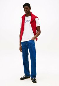 Rød ærmeløs strik over en hvid T-shirt med blå skrift, parret med blå jeans. Model iført sorte sko, stående med den ene hånd i lommen.