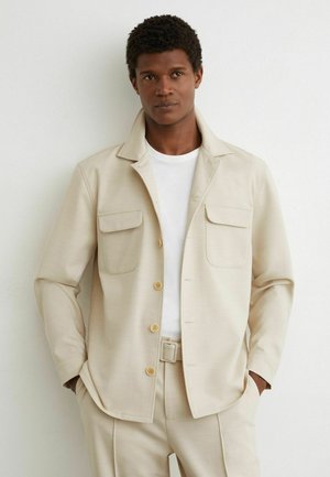 Man die een beige jas met klepzakken draagt over een wit T-shirt en bijpassende beige broek met riem, staand tegen een witte muur.