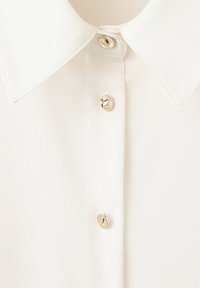 Gros plan d'un col de chemise blanche et d'une patte de boutonnage avec trois boutons dorés brillants visibles, attachés.