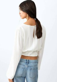 Bílá halenka s dlouhým rukávem a detailem v podobě průstřihu na zádech, texturovaná látka a nařasený pas. V kombinaci se světle modrými džínami.