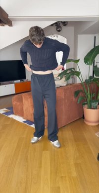 Pantalones negros, holgados, con una cintura elástica. La persona está subiendo los pantalones sobre una camiseta de manga larga azul marino con textura de canalé y unos zapatos grises.