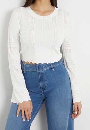 Kobieta nosząca biały, długi rękaw sweterkowy crop top z wzorzystymi detalami, połączony z niebieskimi jeansami z wysokim stanem i pasującym paskiem.