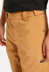 Svetlosivé vodoodpudivé nohavice s hladkým povrchom, plochým predným dielom a dvojitým gombíkom. Obsahujú bočnú kapsu a logo na nohe.