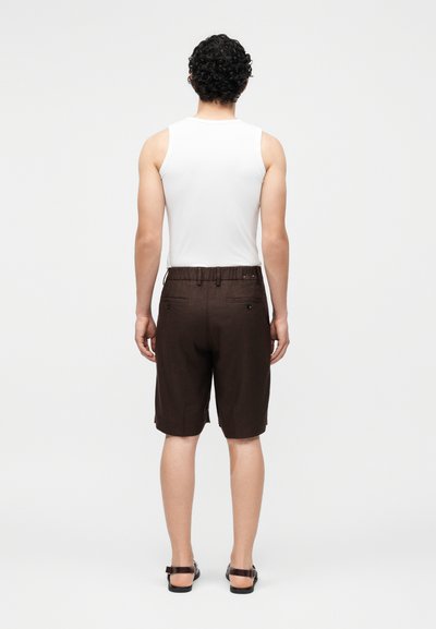 Άτομο με σγουρά μαλλιά που φοράει λευκό αμάνικο μπλουζάκι, καφέ σορτς και μαύρα σανδάλια, στέκεται με την πλάτη σε μονόχρωμο φόντο.