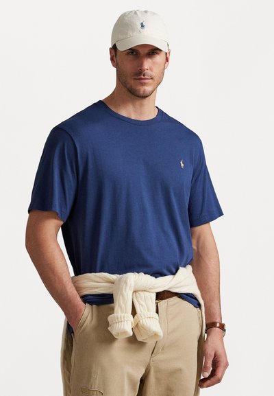 Marineblaue Baumwoll-T-Shirt mit kurzen Ärmeln, das ein kleines Logo zeigt. Cremefarbener Strickpullover um die Taille gebunden. Beigefarbene Hose und Gürtel.