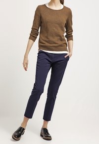 Maglione a maglia marrone con scollatura rotonda indossato su una camicia bianca, abbinato a pantaloni blu navy alla caviglia e scarpe nere con lacci.