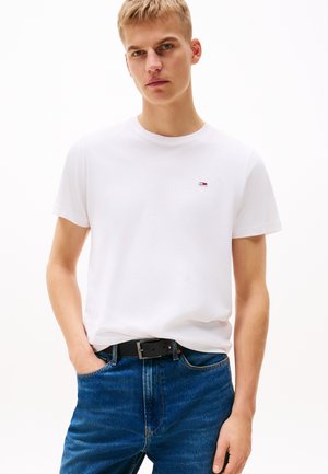 Bela bombažna majica z okroglim izrezom, na prsih manjša večbarvna nalepka. Kombinirana z modrimi jeans hlačami in črnim pasom.