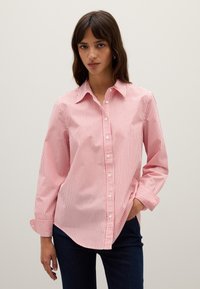 Donna con lunghi capelli castani, che indossa una camicia a righe verticali rossa e bianca e pantaloni blu scuri, con una mano in tasca.