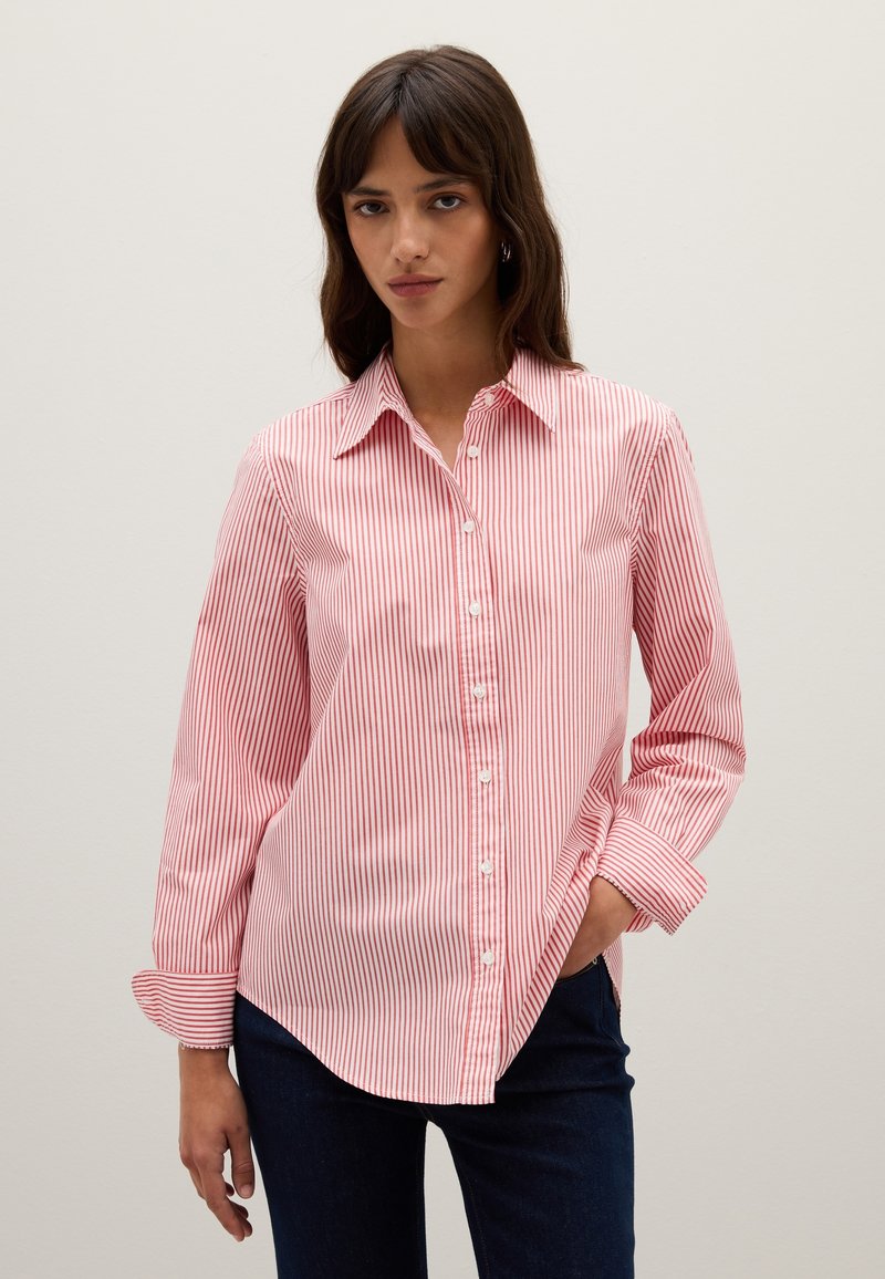 Donna con lunghi capelli castani, che indossa una camicia a righe verticali rossa e bianca e pantaloni blu scuri, con una mano in tasca.