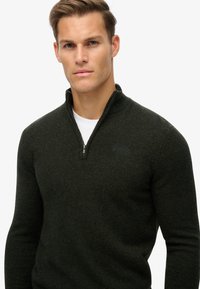 Maglione zip-up verde scuro con colletto alto. Tessuto a maglia morbido e strutturato. Presenta una zip in metallo e un logo discreto sul petto.