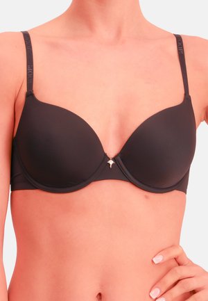 Nainen pukeutunut mustaan muotoiltuun rintaliiveihin, joissa on ohuet olkaimet ja pieni koristeellinen riipus keskellä edessä.