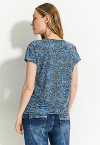 T-shirt grigio con un motivo astratto blu, maniche corte, scollatura rotonda e vestibilità rilassata, indossato con jeans blu.