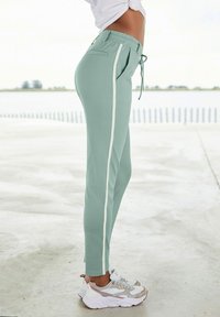 Hellgrüne Sweatpants mit weißen Seitenstreifen, elastischem Bund und Taschen, kombiniert mit weißen, grauen und rosa Sneakern auf einem schlichten Boden.