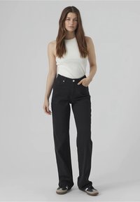 Donna con lunghi capelli castani che indossa una maglietta senza maniche bianca, jeans neri a vita alta e sneakers bianco e nere, in piedi contro uno sfondo semplice.