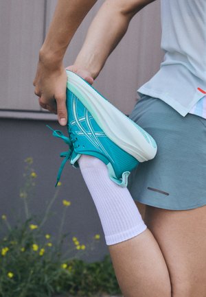 Scarpa sportiva turchese con superficie texturizzata, lacci e suola bianchi. Persona che si estende una gamba indossa un calzino bianco e pantaloni sportivi grigi.