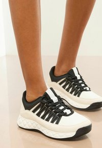 Adidași alb-negru cu un strat texturat, talpă groasă și un design de șireturi încrucișate. Modelele circulare de-a lungul tălpii adaugă detalii.