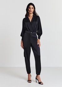 Blusa nera in satin con scollo a V e maniche a sbuffo, abbinata a pantaloni neri con polsini elastici e chiusura a bottone. Scarpe con tacco alto.