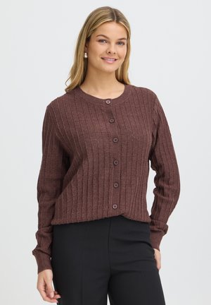 Femme aux longs cheveux blonds porte un cardigan marron côtelé à boutons et un pantalon noir, debout devant un fond clair uni, souriante.