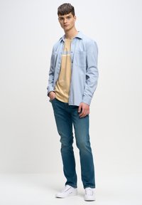 Camisa abotonada azul clara sobre una camiseta beige con una raya blanca y azul, combinada con jeans azules y zapatillas blancas.