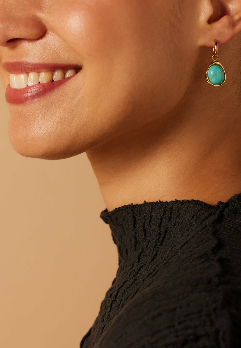 Boucles d'oreilles pendantes dorées avec des pierres turquoise en forme de goutte, présentant un motif marbré, portées avec un haut noir texturé.