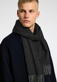 Sciarpa a zigzag grigio scuro con estremità frangiate, realizzata in un tessuto morbido. Indossata attorno al collo, abbinata a un cappotto blu navy.