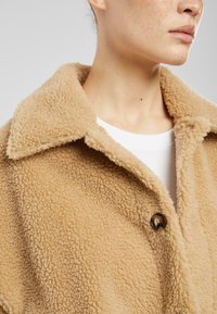 Giacca in faux shearling color ocra con grande colletto, caratterizzata da un singolo bottone marrone e un tessuto morbido e testurizzato.