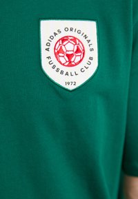 Žalias medvilninis marškinėliai su baltu šarvo logotipu, kuriame pavaizduota raudona futbolo kamuolio dizainas ir tekstas: "ADIDAS ORIGINALS FUSSBALL CLUB 1972."