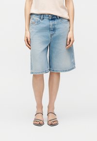 Γυναίκα που φοράει ανοιχτό μπλε τζιν σορτσάκι μέχρι το γόνατο, μπεζ τοπ και μαύρα σανδάλια με λουράκια σε λευκό φόντο.