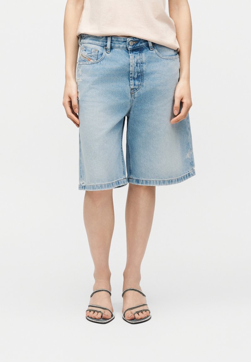 Γυναίκα που φοράει ανοιχτό μπλε τζιν σορτσάκι μέχρι το γόνατο, μπεζ τοπ και μαύρα σανδάλια με λουράκια σε λευκό φόντο.