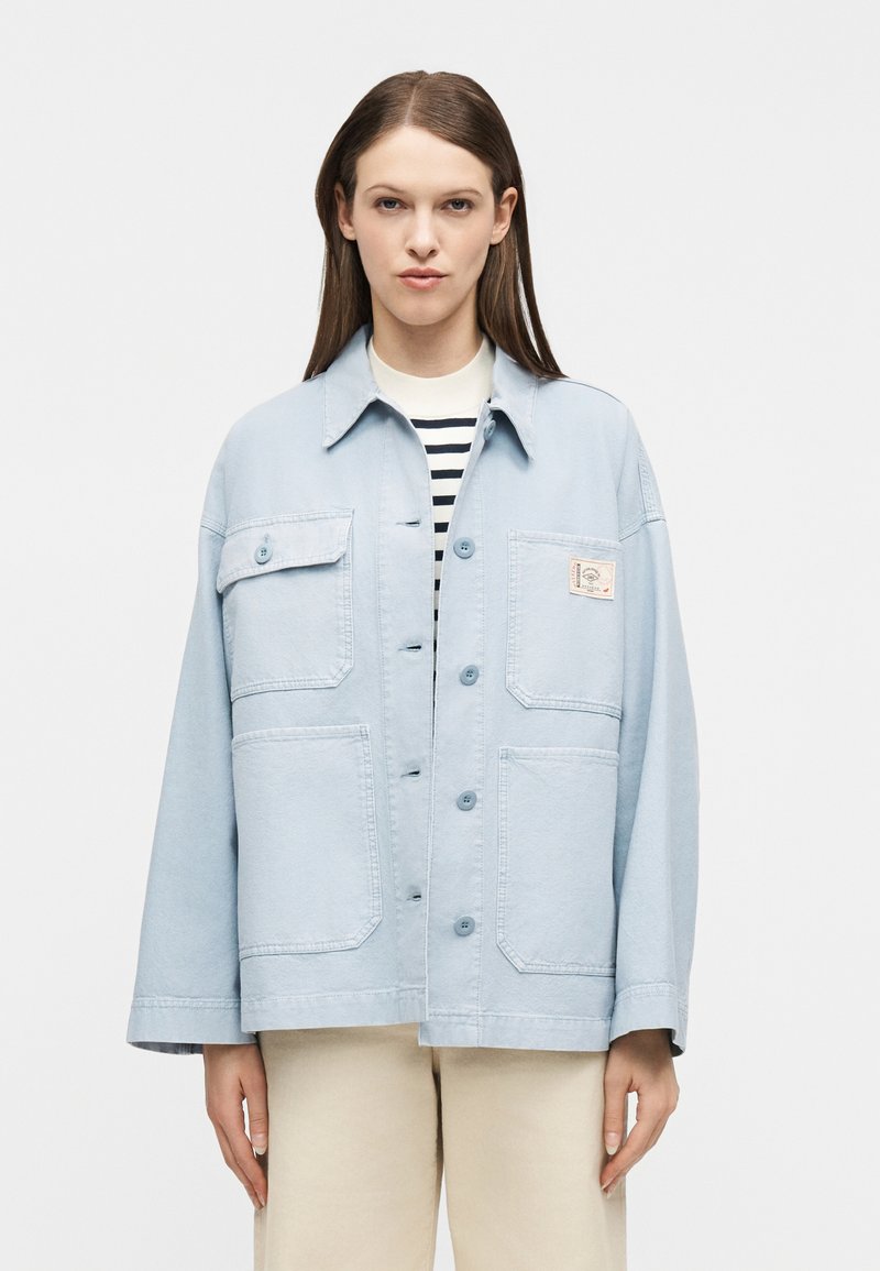 Γυναίκα που φοράει ανοιχτό μπλε υπ ερμεγέθη σακάκι με κουμπιά και μεγάλες τσέπες πάνω από ριγέ πουκάμισο και μπεζ παντελόνι, που στέκεται μπροστά σε λευκό φόντο.