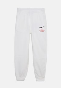 White sweatpants with an elastic waistband, side pockets, black Nike logo, and red text detail on the left leg. Cuffed ankles.
