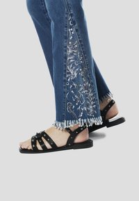 Farmernadrág fehér és ezüst hímzett virágmintákkal az oldalán; foszlott szegéllyel. Fekete pántos szandál ezüst díszítéssel.