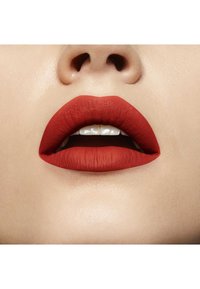 Rouge à lèvres mat de couleur rouge appliqué sur des lèvres pulpeuses, mettant en valeur une texture lisse avec un léger éclat. Dents visibles, fond doux et neutre.