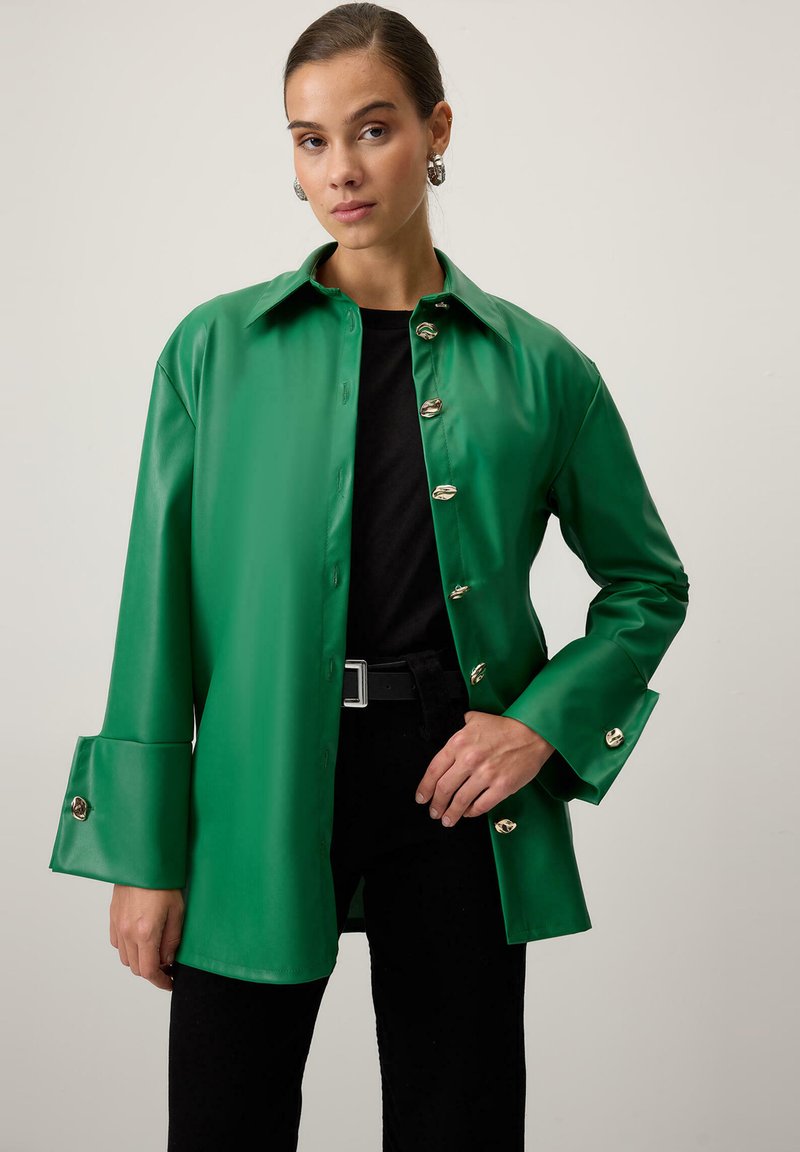 Zaļš mākslīgās ādas krekla jaka ar apkakli, metāla pogu detaļām un platām piedurknēm. Kombinēta ar melnu krekla un biksēm.