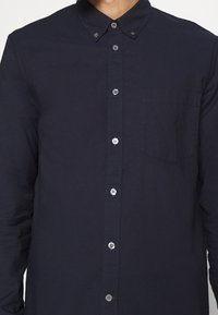 Homme portant une chemise bleu marine à manches longues boutonnée, avec une poche poitrine et un col boutonné, visible du cou au milieu du torse.