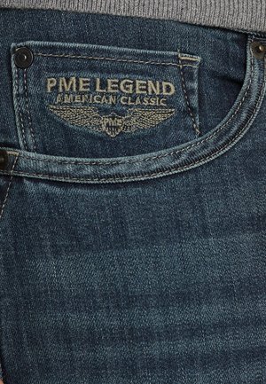 Plave traperice s mornarskim pojasom. Stražnji džep prikazuje "PME Legend" logo u bež koncu, s krilatim simbolom.