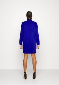 Blå stickad klänning med långa ärmar och hög krage. Har elastisk fåll och mudd. Bärs med svarta klacksandaler.