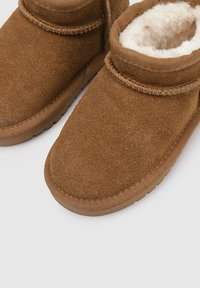 Botas de tobillo de ante marrón con forro de felpa crema, punta redonda y costuras mínimas. Textura suave y suela de goma flexible para mayor comodidad.