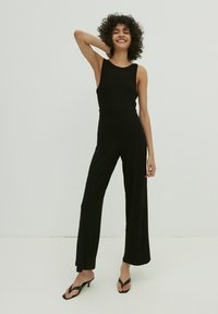 Schwarzer, gerippter Jumpsuit mit ärmellosem Design, tailliertem Oberteil und weitem Bein. Das Model kombiniert ihn mit schwarzen Flip-Flop-Sandalen.