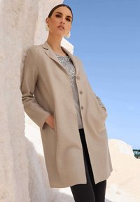 Beige Wollmantel mit Reverskragen, vorderen Knöpfen und Seitentaschen, getragen über einem grauen Oberteil mit Pailletten. Das Model steht vor einem hellen Hintergrund.