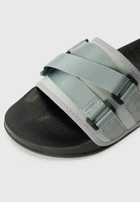 Sandal na klizanje s teksturiranom crnom podlogom i širokom trakom u svijetlo plavoj boji, s dizajnom križnih traka i sivi akcentima. Izrađeno od izdržljivog materijala.