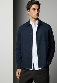 Navy woven shirt jacket with a collar, front button closure, and one chest pocket, layered over a white collared shirt.