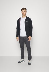 Svart zip-up jakke med mønstret innside, grå t-skjorte, grå bukser og fargerike joggesko. Modellen står mot en nøytral bakgrunn.