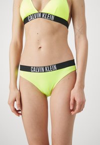 Neon gele bikini set met een driehoekige bralette en bijpassende slipjes, beiden met een zwarte Calvin Klein logo tailleband. Gladde textuur.