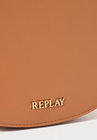 Lærveske i tan med en glatt tekstur og gull "REPLAY" logo. Rund form med sømmer i kantene. Minimalistisk design uten ekstra detaljer.